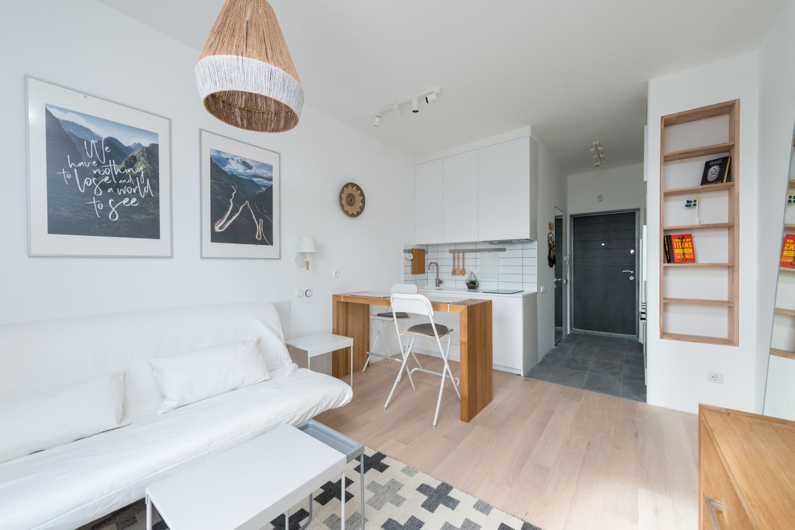 Studio pequeno e claro com sofá branco, tapete geométrico e piso de madeira clara, ambiente integrado