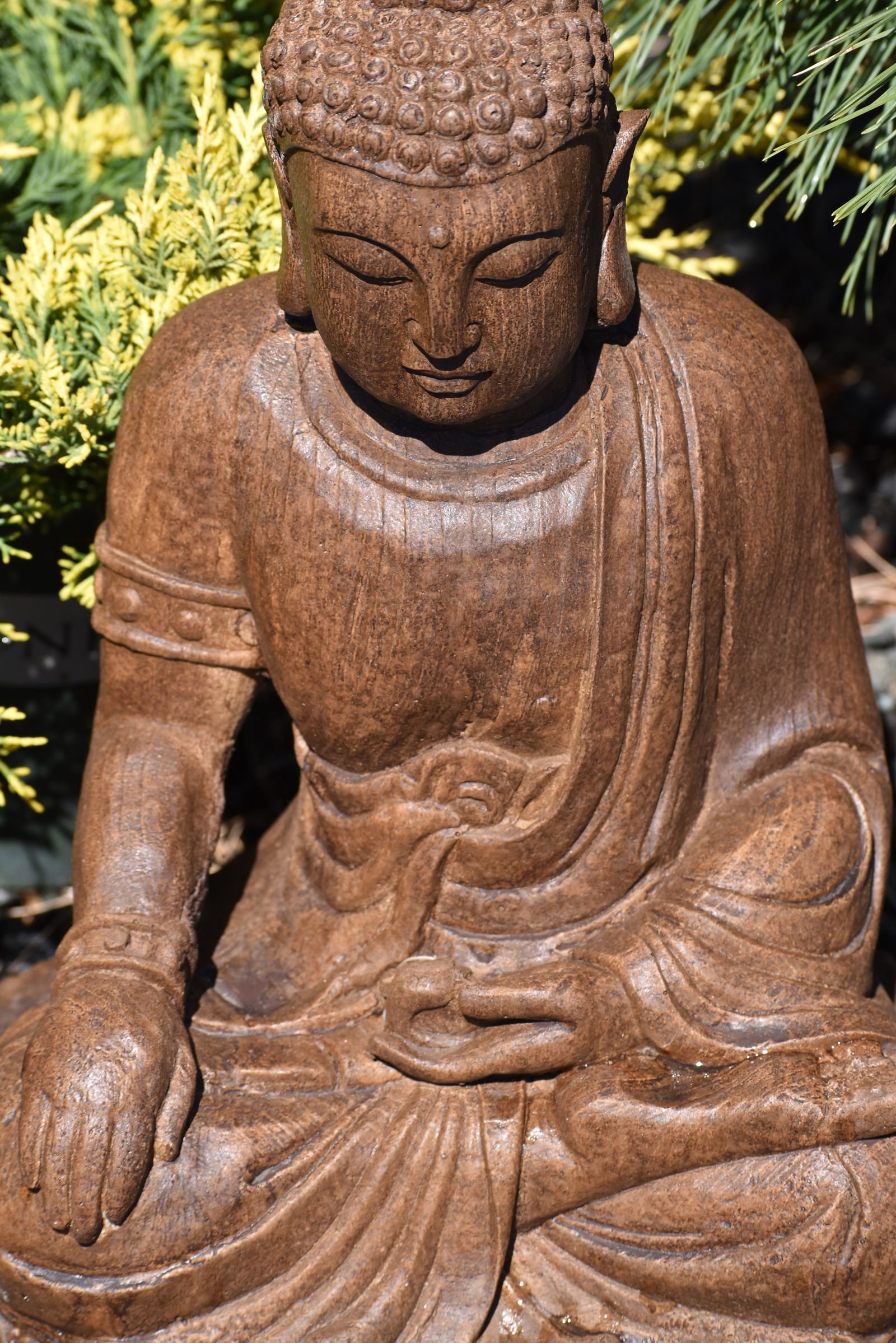 Estátua de Buda sentado em meditação, esculpida em pedra marrom, em meio a folhagem verde de jardim