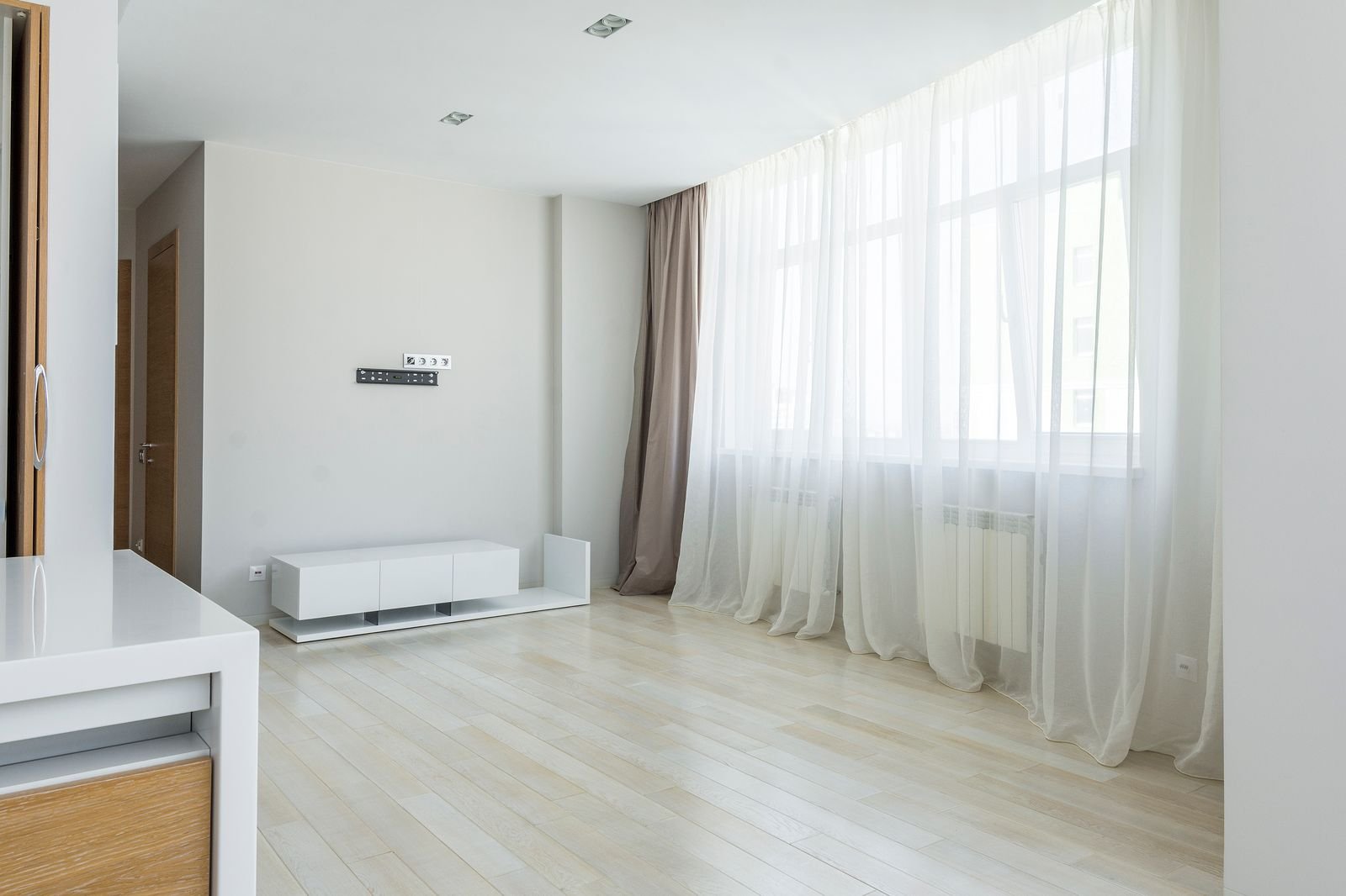 Sala clara e vazia com cortina de voil branca em janela ampla, piso de madeira clara e ambiente arejado