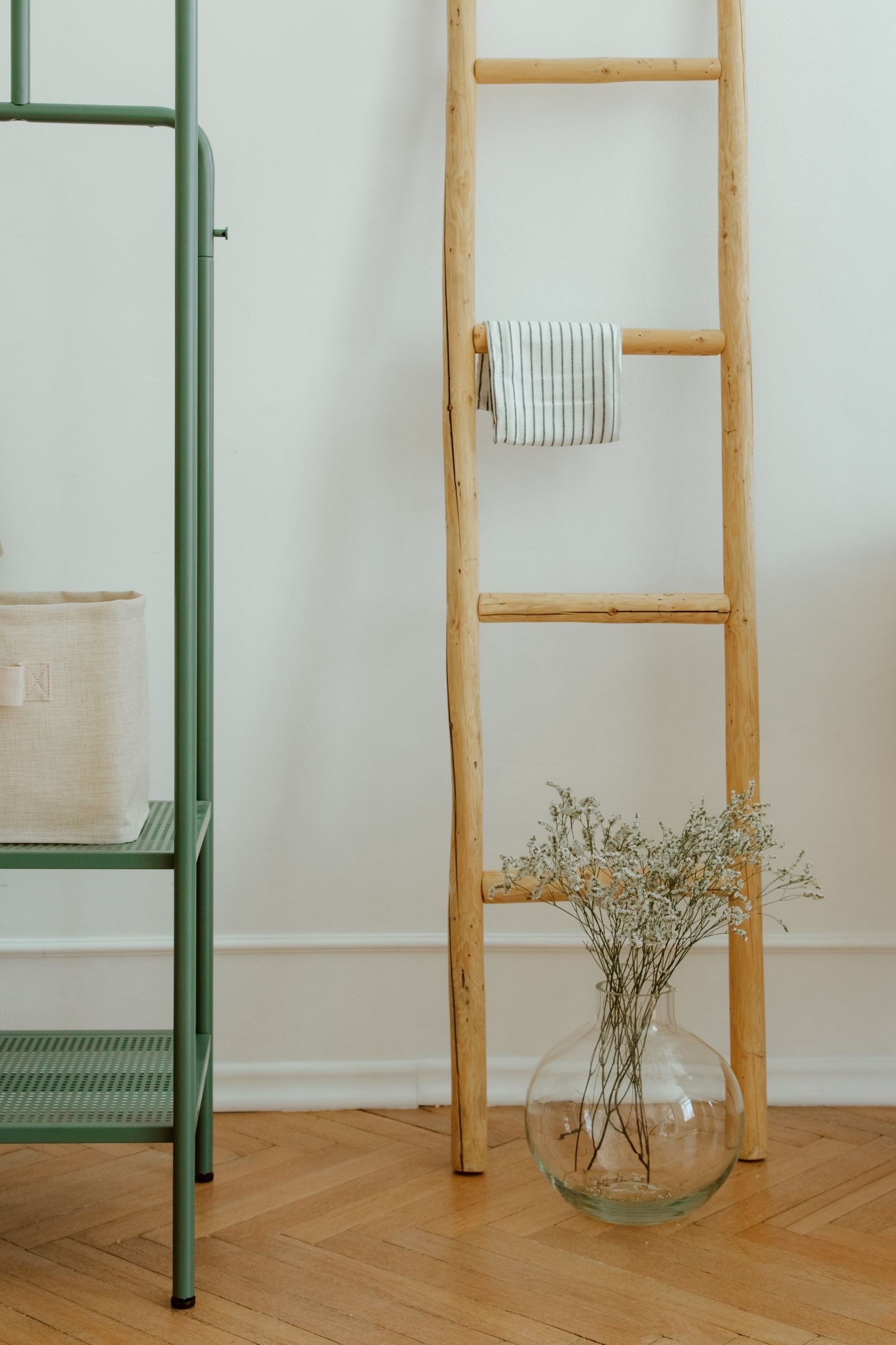 Canto de casa organizado com estante verde clara, cesto de tecido, escada de madeira com pano listrado e vaso de vidro com flores secas
