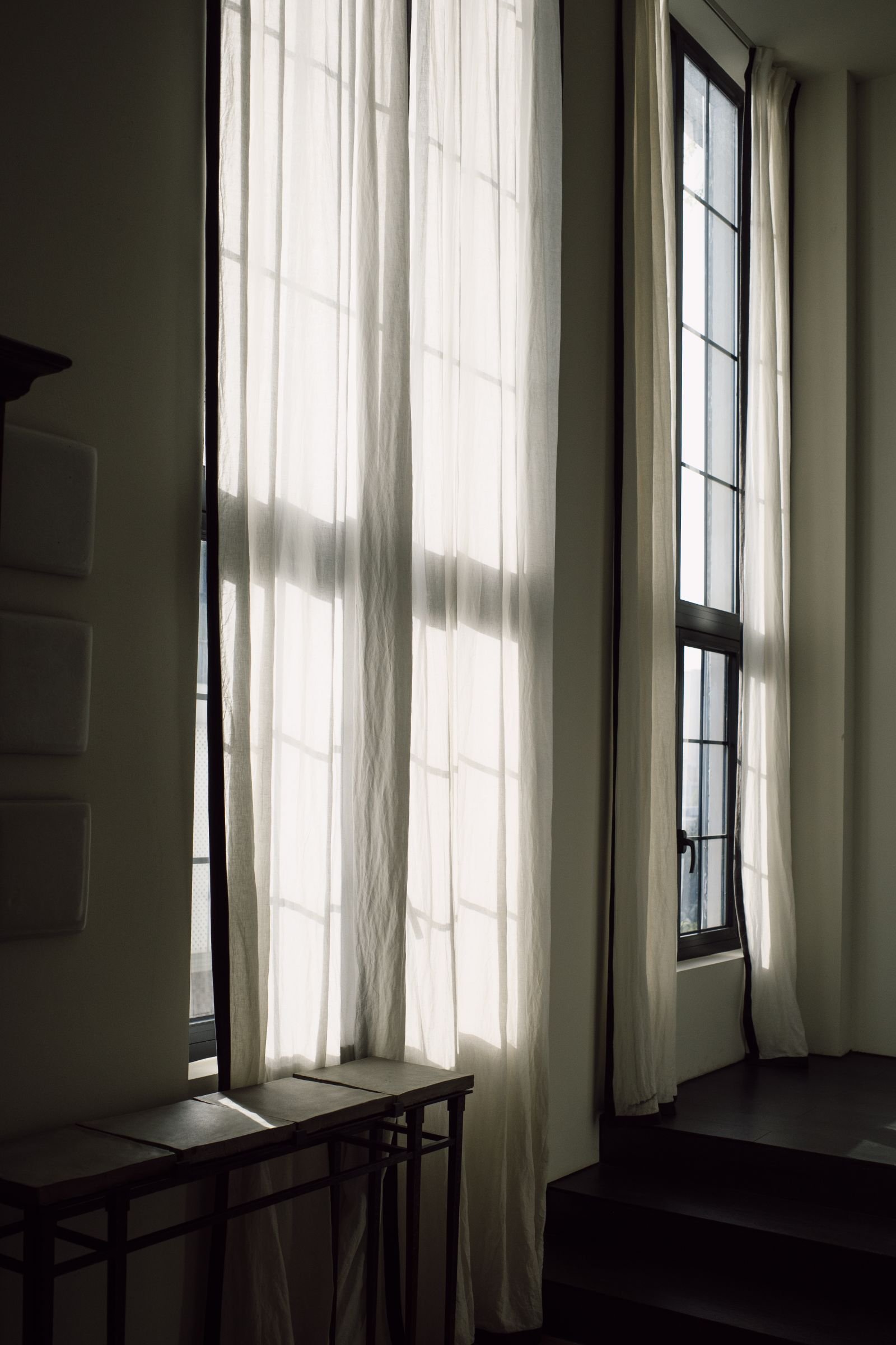 Duas janelas altas com cortinas de linho claro, cada uma deixando entrar luz de um ângulo diferente