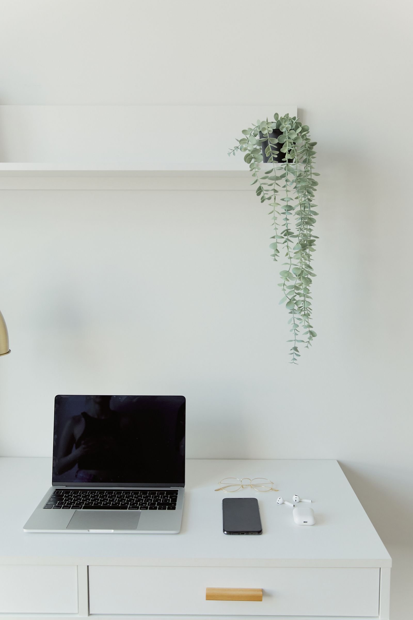 Mesa de trabalho minimalista branca, com notebook, óculos, fones sem fio e planta pendente na parede