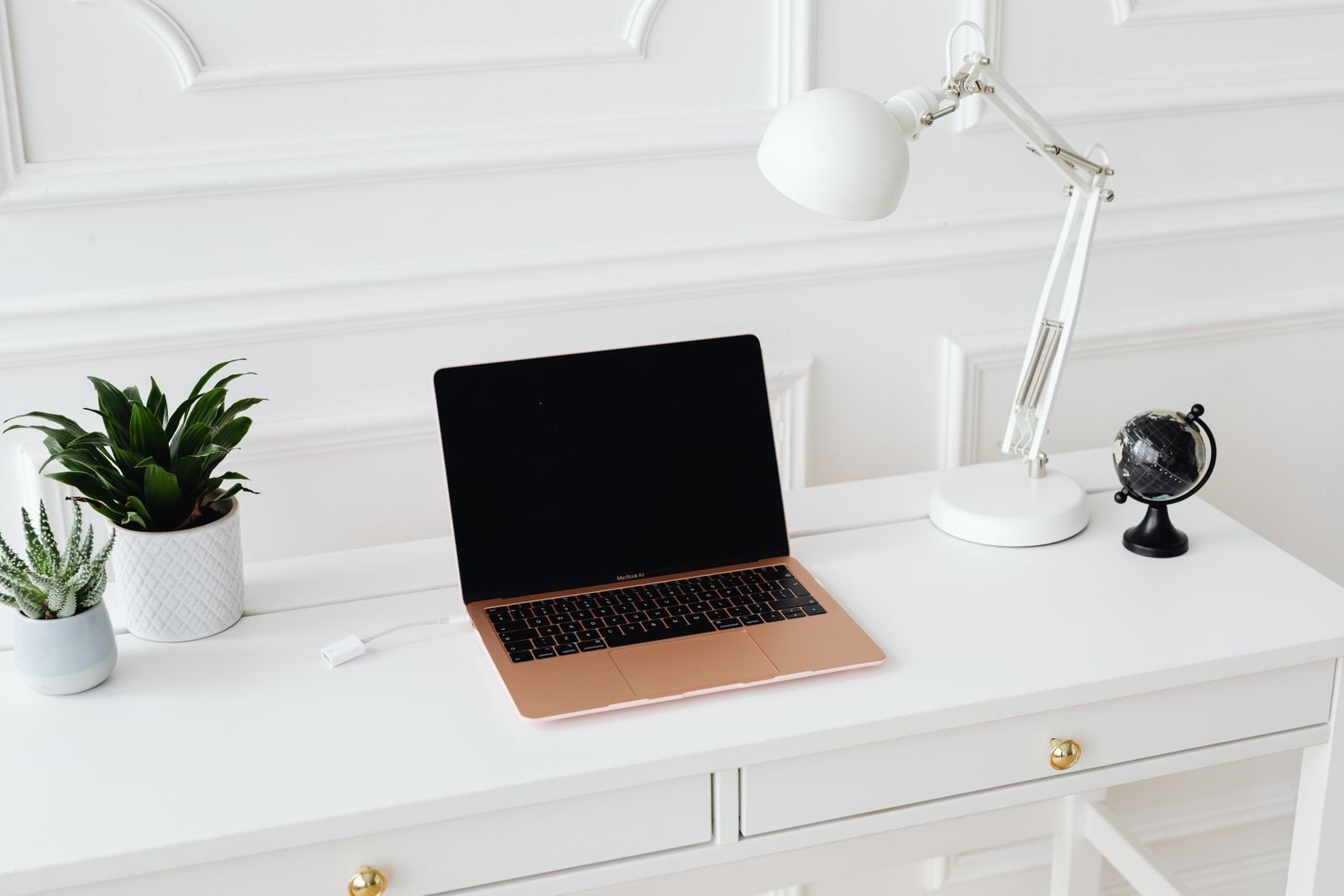 Mesa de trabalho branca e elegante, com notebook, luminária articulada, plantas e globo terrestre