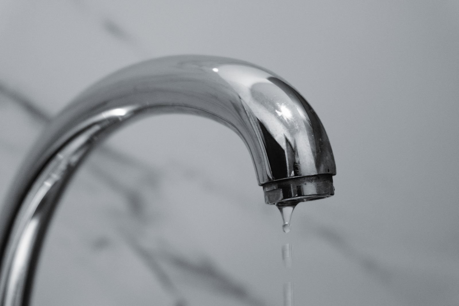 Torneira cromada curva com uma gota de água escorrendo do bico, contra fundo de mármore claro