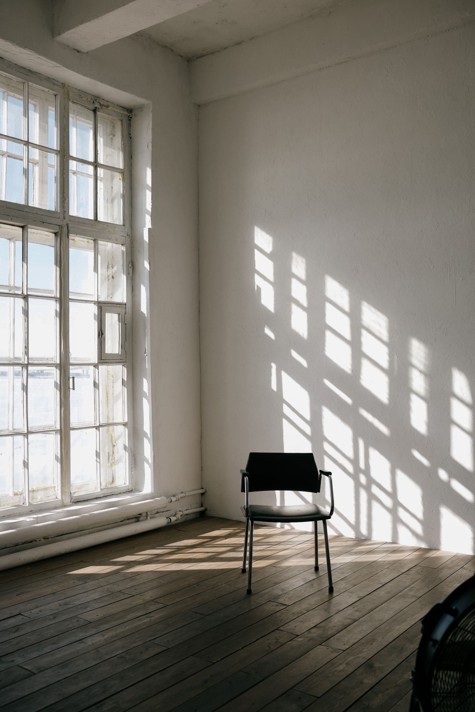 Sala ampla com janela grande, luz forte entrando e sombras da grade desenhadas na parede e no piso de madeira